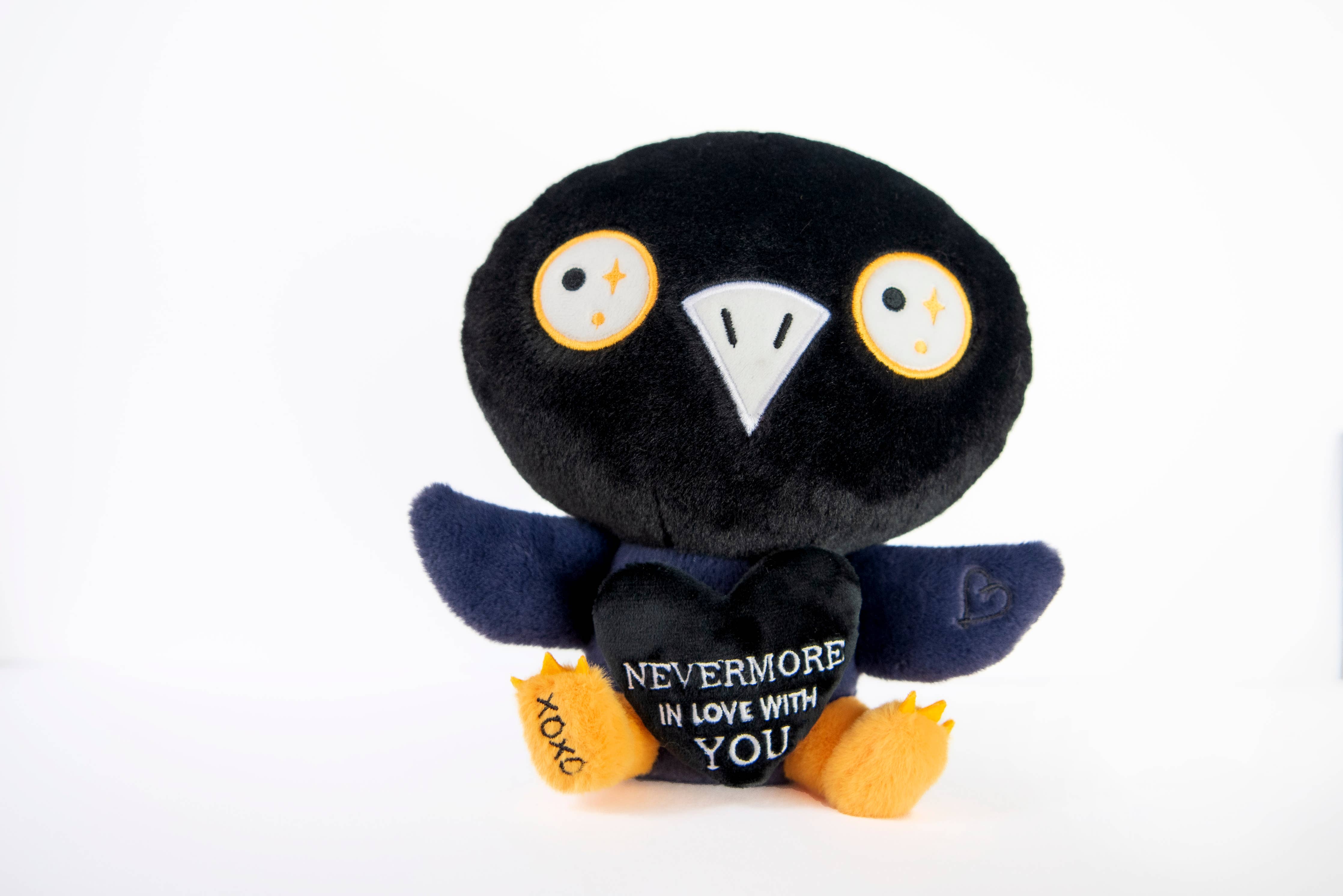 A black and blue raven plushie with a white beak and white and yellow eyes. Its wings are holding a black heart with the white text "Nevermore in love with you." The raven has yellow legs with a black "xoxo" on the right foot.