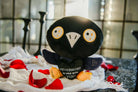 A black and blue raven plushie with a white beak and white and yellow eyes. Its wings are holding a black heart with the white text "Nevermore in love with you." The raven has yellow legs with a black "xoxo" on the right foot.