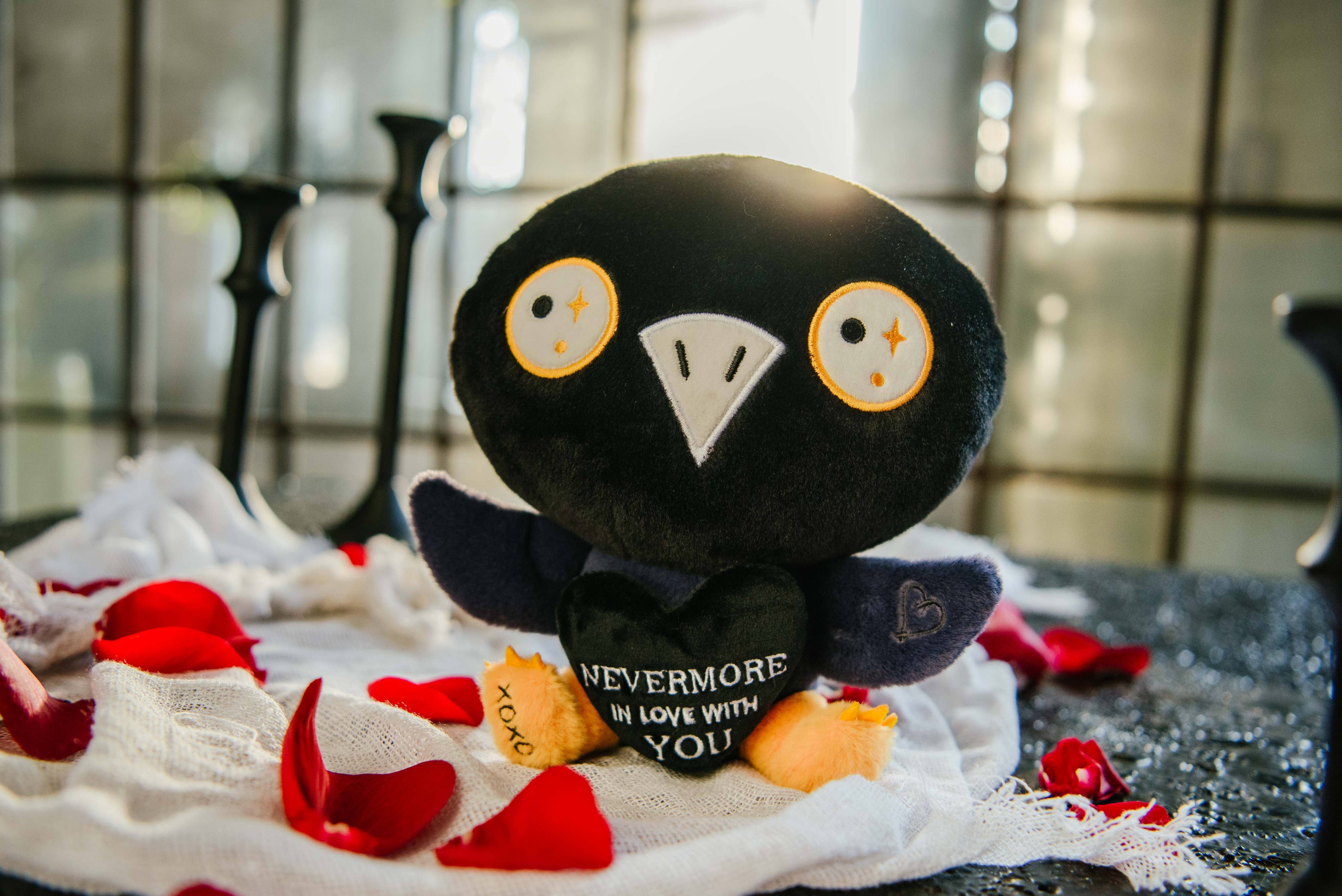 A black and blue raven plushie with a white beak and white and yellow eyes. Its wings are holding a black heart with the white text "Nevermore in love with you." The raven has yellow legs with a black "xoxo" on the right foot.