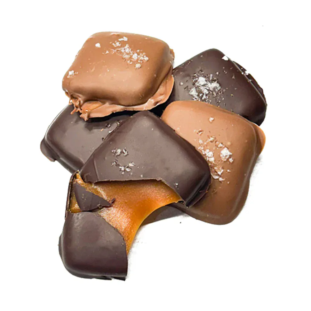 A close-up shot of several gourmet sea salt caramels, with one broken open to reveal a thick, flowing golden caramel center. The chocolates are topped with visible flakes of sea salt and set against a clean white background.