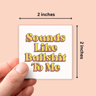 A hand holds a small square white sticker against a peach background. The sticker has retro-style yellow and pink text reading 'Sounds Like Bullshit To Me.