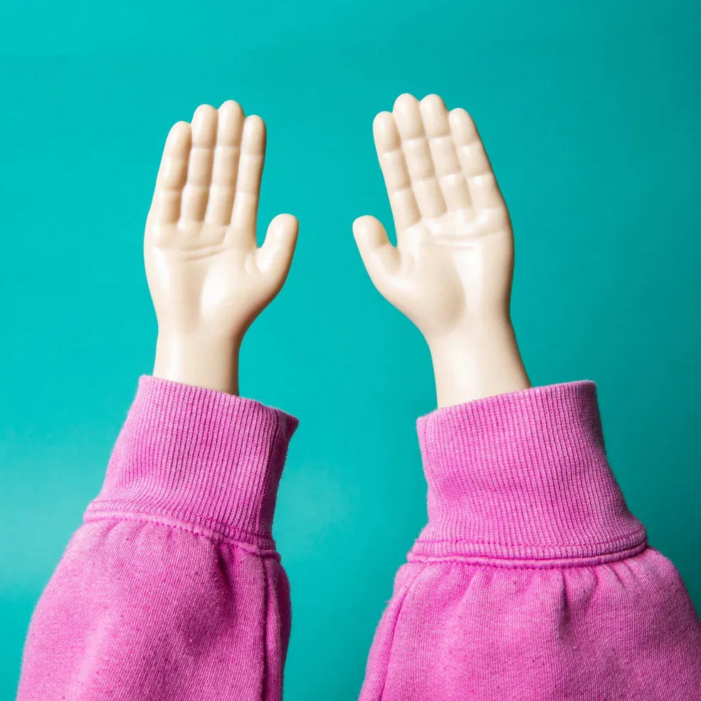 Two plastic hands in the air, wearing pink fabric cuffs, against a turquoise background. The image conveys a playful and whimsical tone.