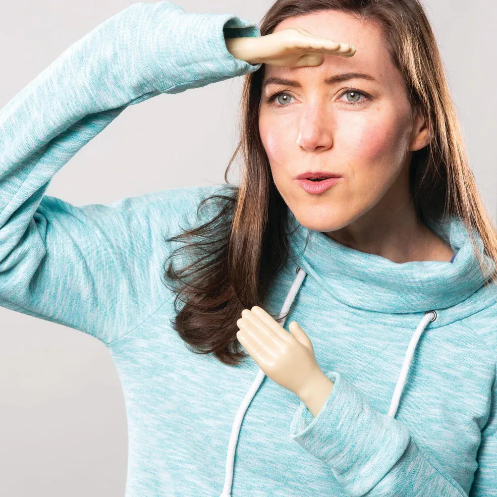 A woman in a teal sweater playfully shields her eyes and mouth with tiny plastic hands attached to her cuffs, conveying curiosity and humor.