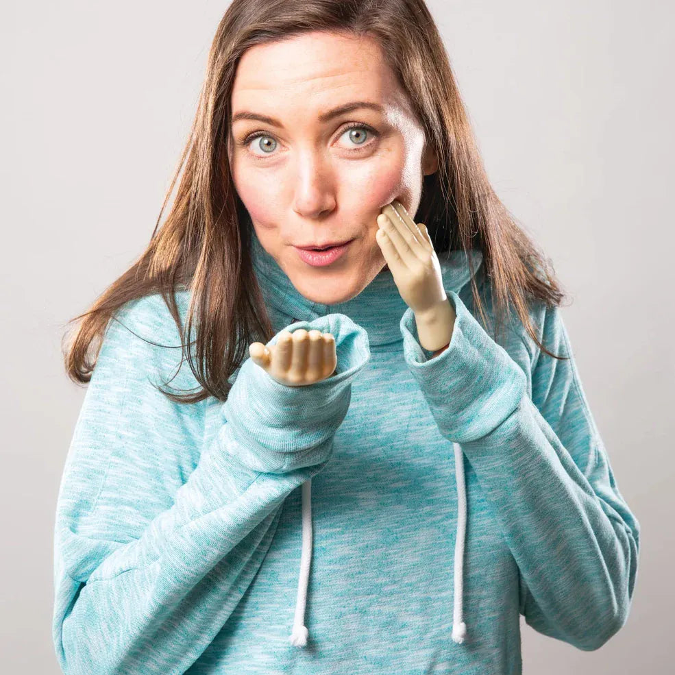 A woman in a teal hoodie playfully uses tiny plastic hands on her fingers, creating a humorous expression. She has a surprised look, enhancing the fun mood.