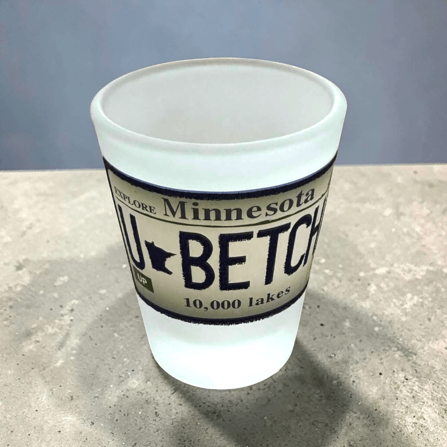 shot glass with Minnesota-themed text and design on a white background. Design looks like a Minnesota License Plate that states "U Betcha"