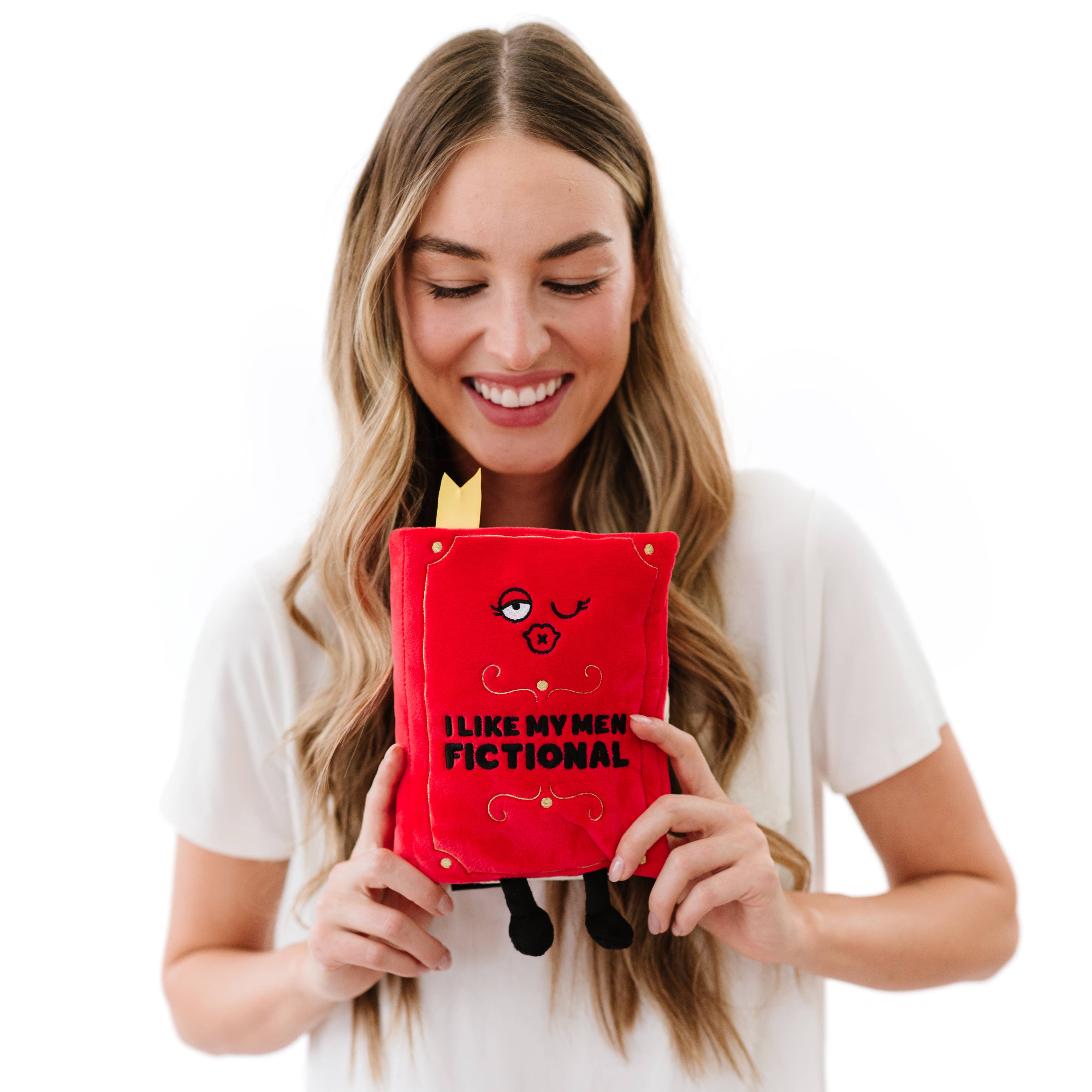 A red plush book with a kissy winky face and embroidered black text that reads 'I like my men Fictional.' The plush has black legs sticking out, a yellow bookmark peeking from the top, and 'A Spicy Romance' embroidered in black on the book's spine. A woman with long blonde hair, wearing a white shirt, is holding the plush.