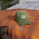 A green cap with "kinda OUTDOORSY" lies on an orange plaid blanket. A hiking boot is partially visible, suggesting an outdoor adventure theme.