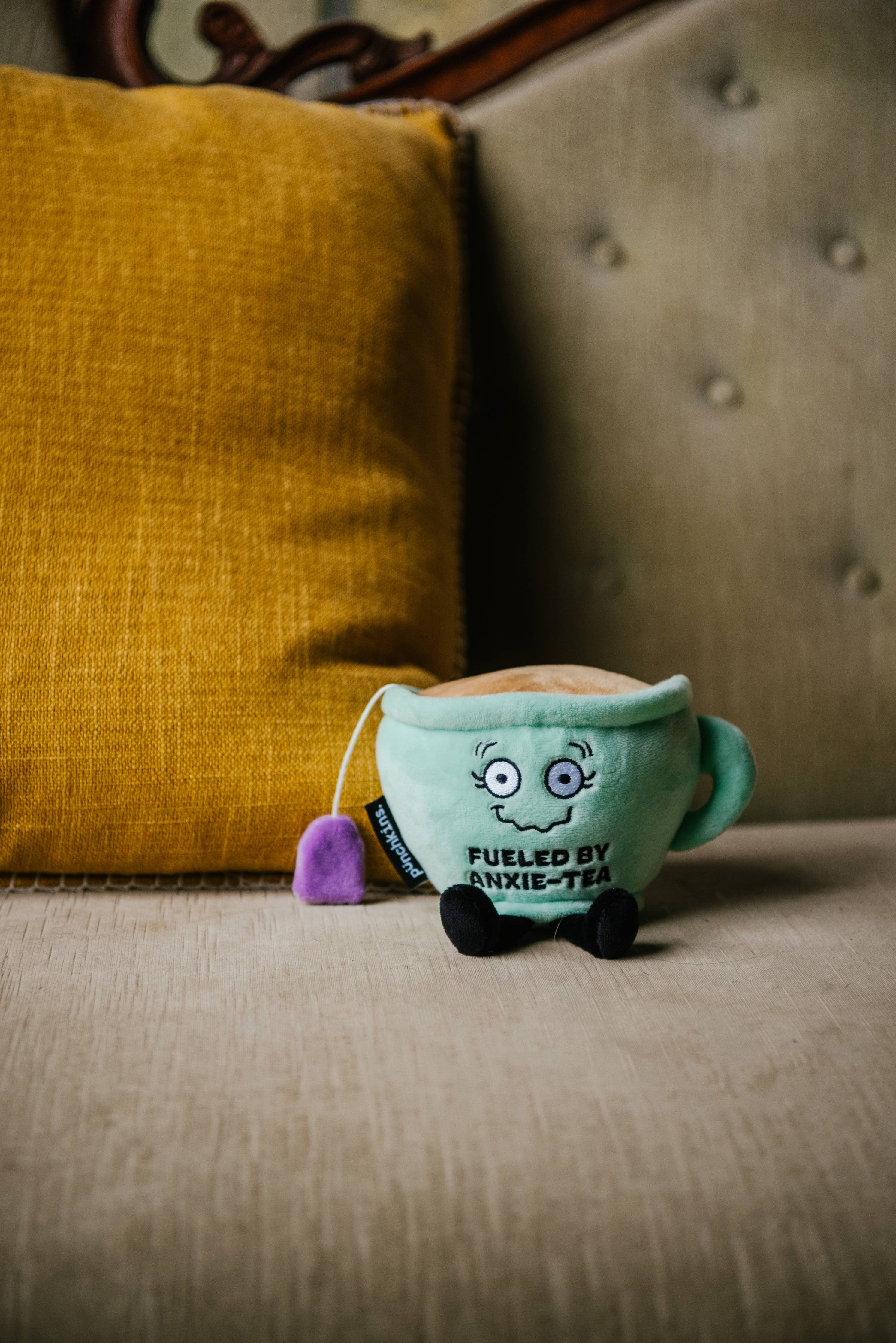 A green plush toy shaped like a smiling tea cup with a shaky smile. The cup is filled with soft, plush 'tea' on top, and a purple tea bag hangs from the side. The front displays the embroidered phrase "FUELED BY ANXIE-TEA" in bold letters. It has small black legs, a handle on one side, and an overall cute, cozy design.