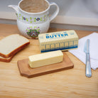Stick of butter on a wooden dish, next to sliced bread, a floral ceramic jug, a butter wrapper, and a knife on a napkin. Cozy kitchen vibe.