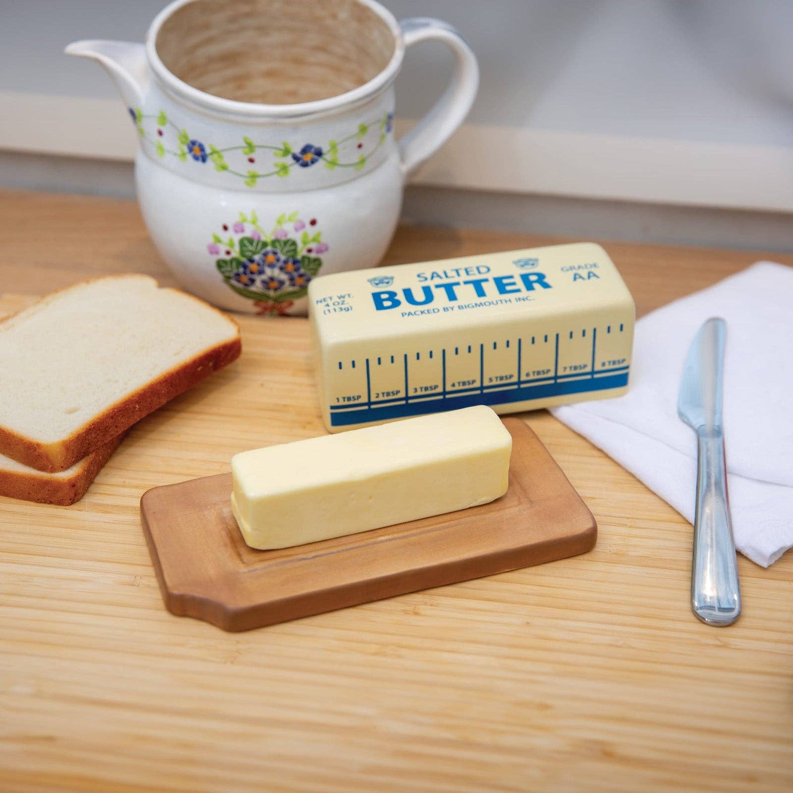 Stick of butter on a wooden dish, next to sliced bread, a floral ceramic jug, a butter wrapper, and a knife on a napkin. Cozy kitchen vibe.