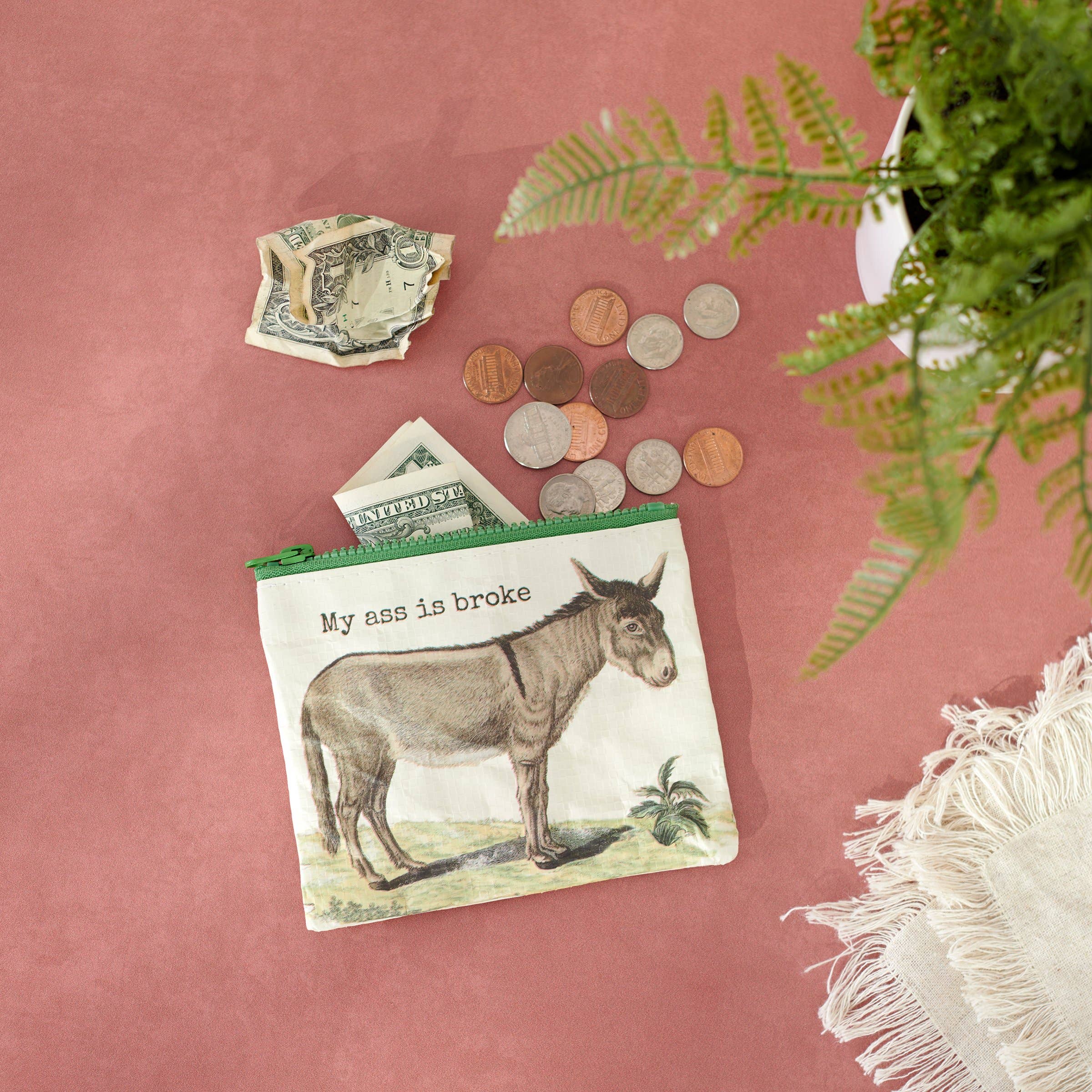 Pouch with a donkey and humorous text saying, "My ass is broke," partially filled with dollar bills, surrounded by coins, a plant, and a tassels on a pink surface.