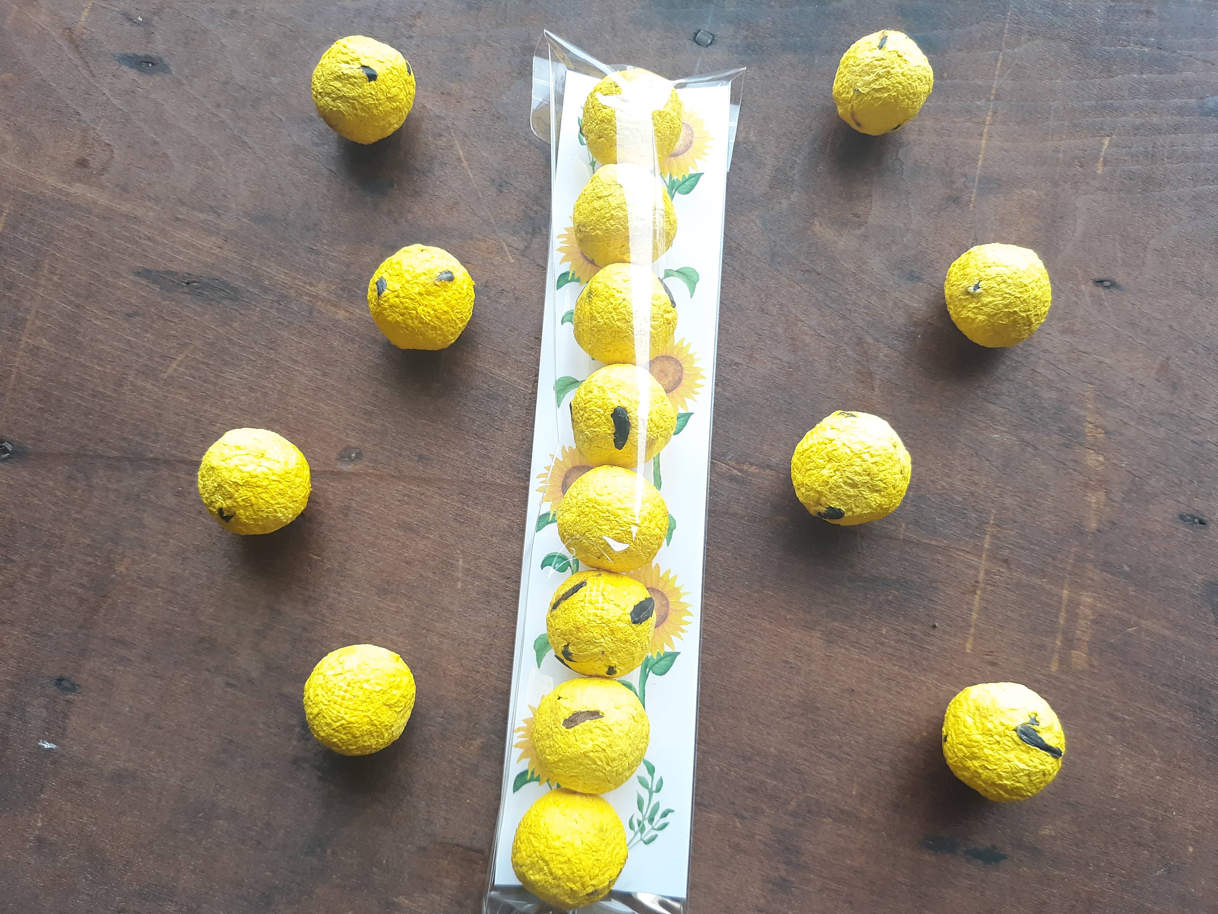 Bright yellow balls resembling lemons, some with painted seeds, are scattered on a wooden surface. A row rests in sealed plastic packaging adorned with sunflower designs.