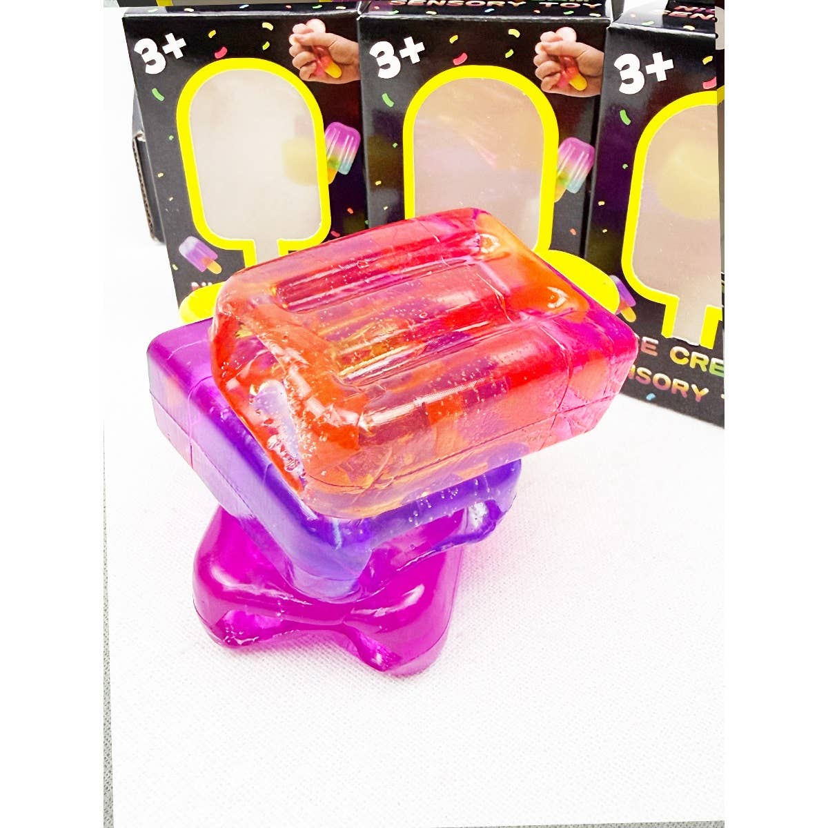 A stack of translucent, rectangle-shaped sensory squishy toys in vibrant pink, orange, and purple, shown in front of their "Ice Cream Bar Squeeze Squishy" retail packaging boxes.
