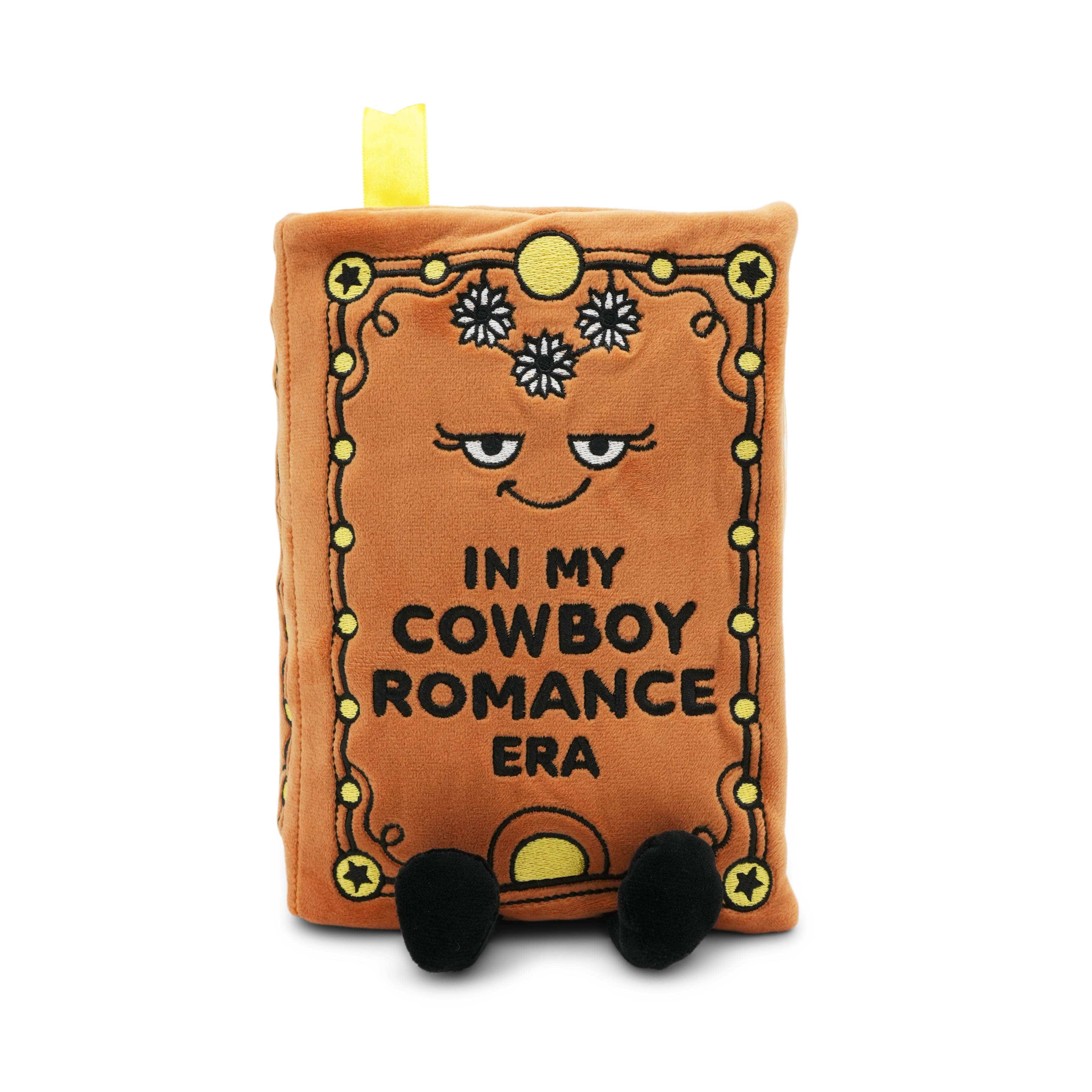 Plush toy resembling a book with a face and text 'In my cowboy romance era' on a white background