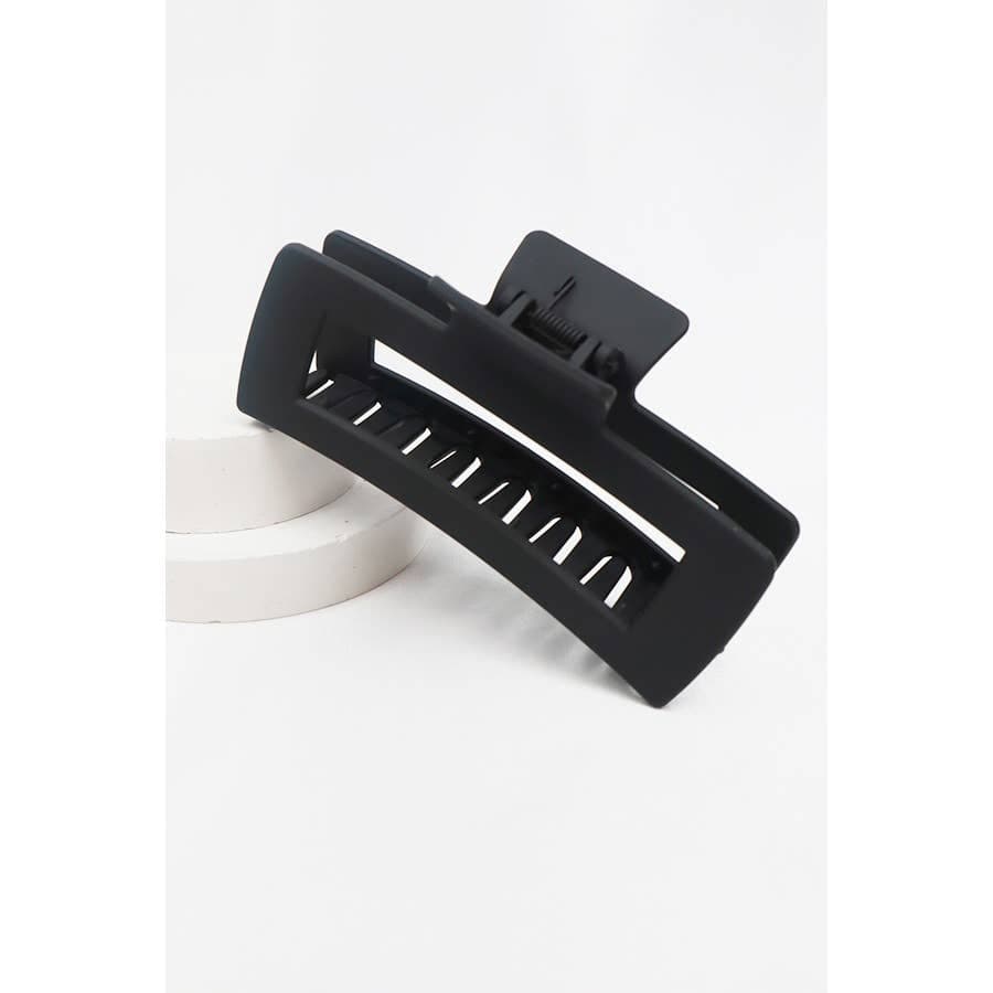A large, matte black jumbo rectangular hair claw clip with a hollow cut-out design, propped on a white circular display platform.