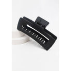 A large, matte black jumbo rectangular hair claw clip with a hollow cut-out design, propped on a white circular display platform.
