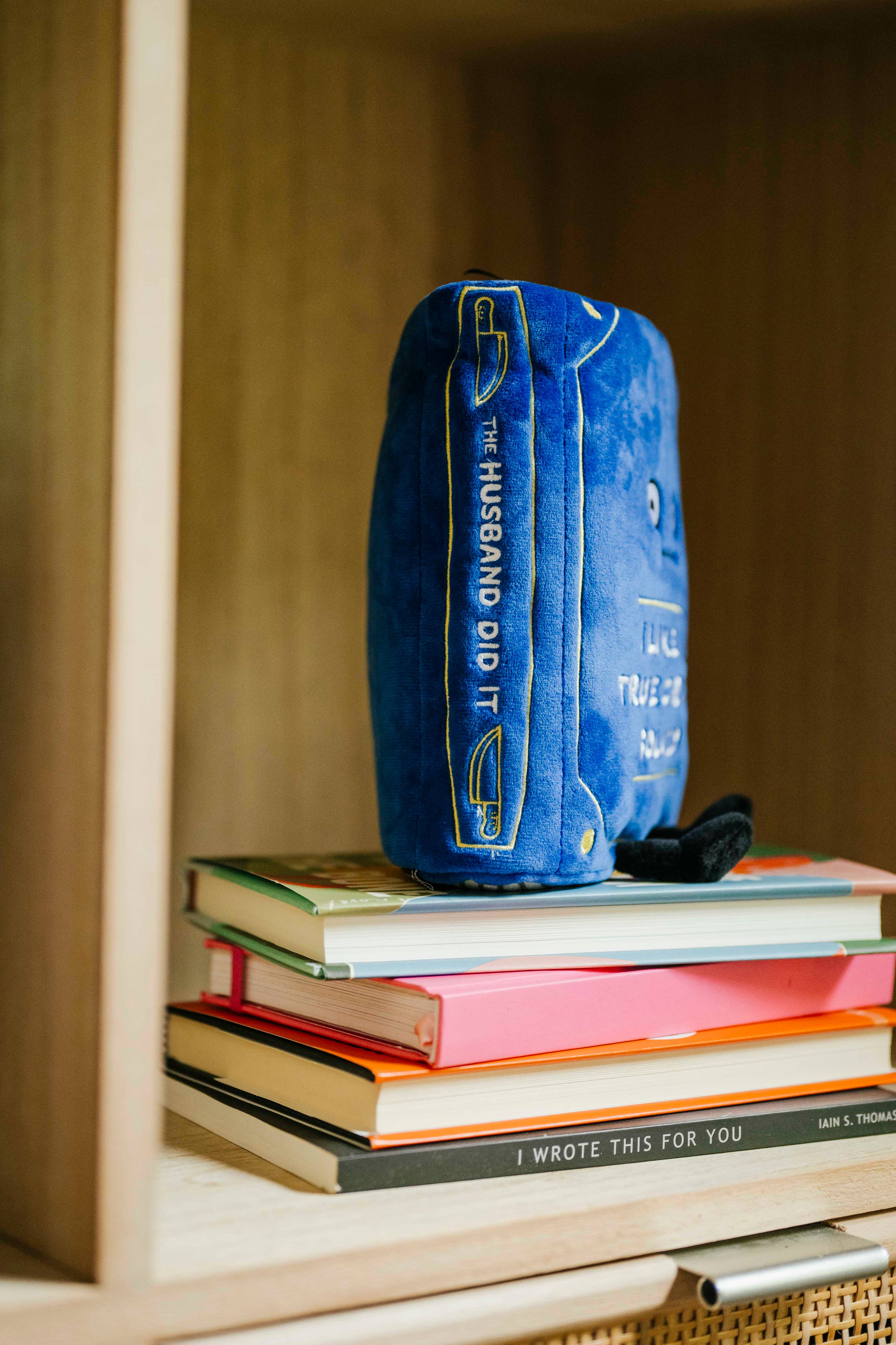 Blue plush book sitting up, with a straight face and white embroidered words that say 'I like my true crimes solved.' Black legs sticking out bottom and black bookmark peeking out the top. Plush is sitting on a bookshelf with the spine facing. Spine has white embroidery that says 'the husband did it'