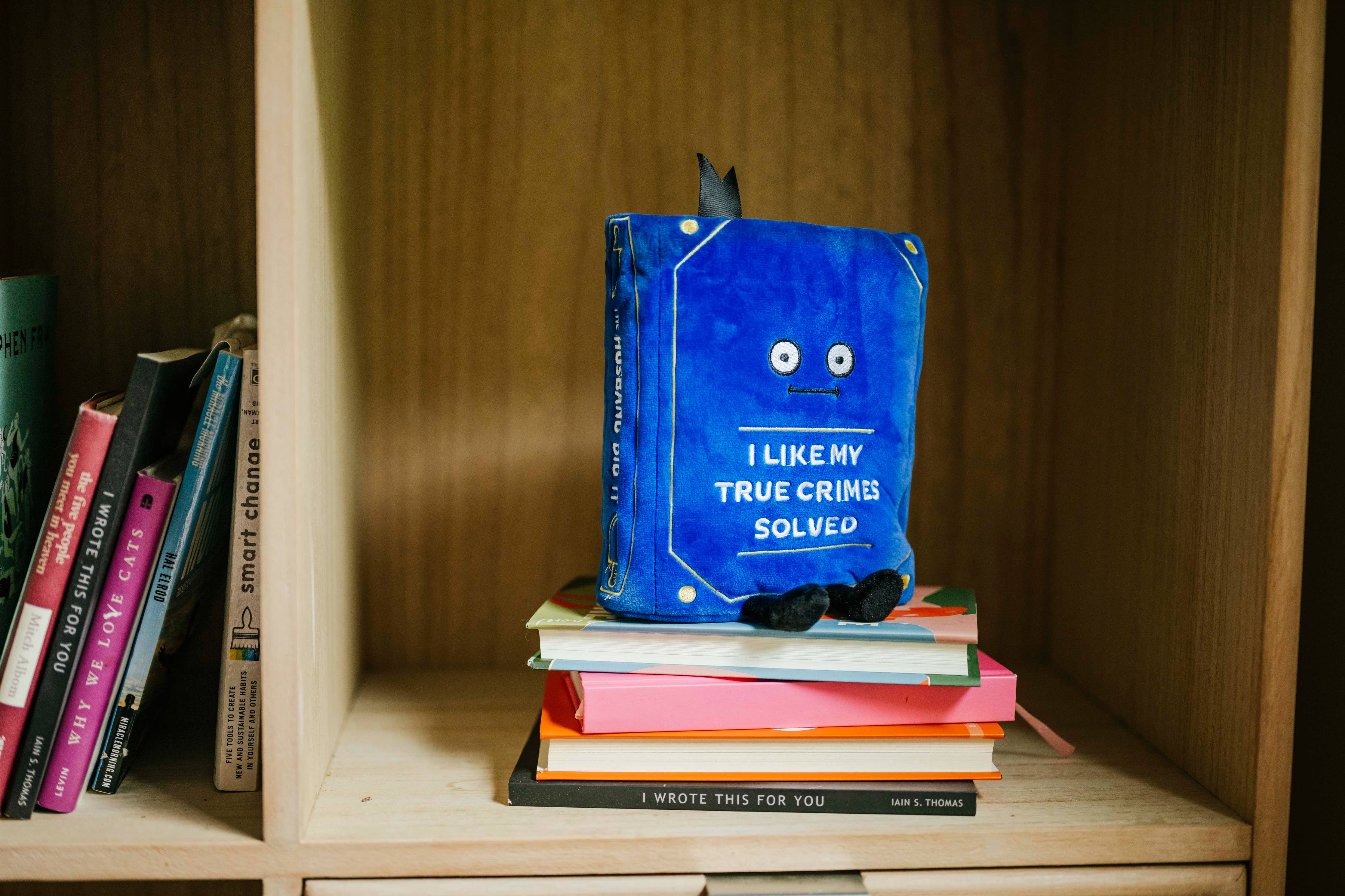 Blue plush book sitting up, with a straight face and white embroidered words that say 'I like my true crimes solved.' Black legs sticking out bottom and black bookmark peeking out the top. Plush is sitting on a bockshelf