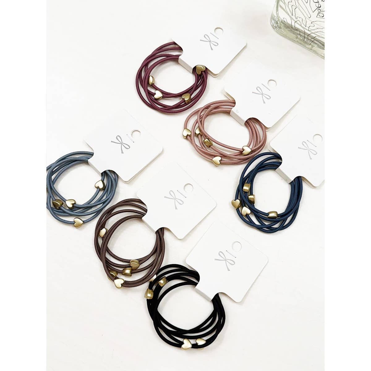 Colorful hair ties with heart-shaped beads are arranged on white cards. Shades include burgundy, pink, blue, brown, and black on a white background.
