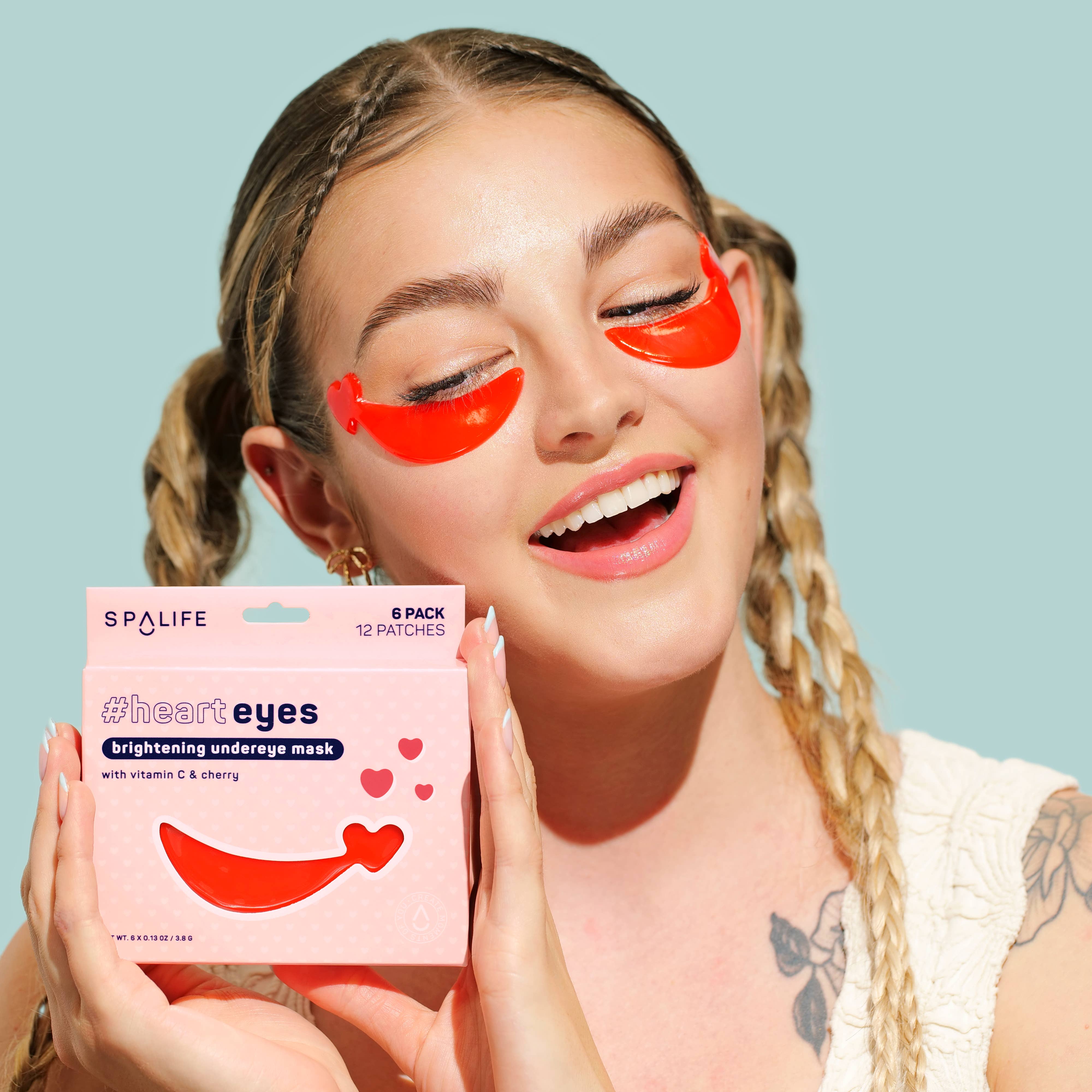 Smiling person with braided hair wears bright red undereye patches, holding SpaLife #heart eyes mask box. Background is light teal. Mood: cheerful.