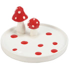 Ceramic plate featuring two red and white spotted mushroom figurines. The white plate is dotted with red spots, conveying a whimsical, decorative tone.