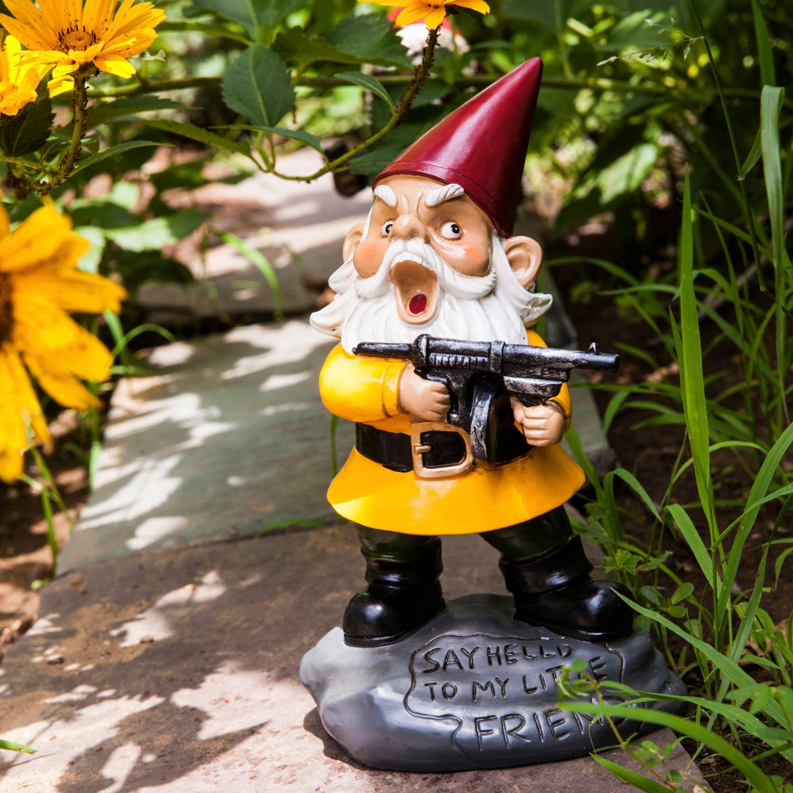 Garden gnome in a yellow coat and red hat holds a toy gun. It stands on a stone with “Say hello to my little friend” engraved, surrounded by greenery and flowers.