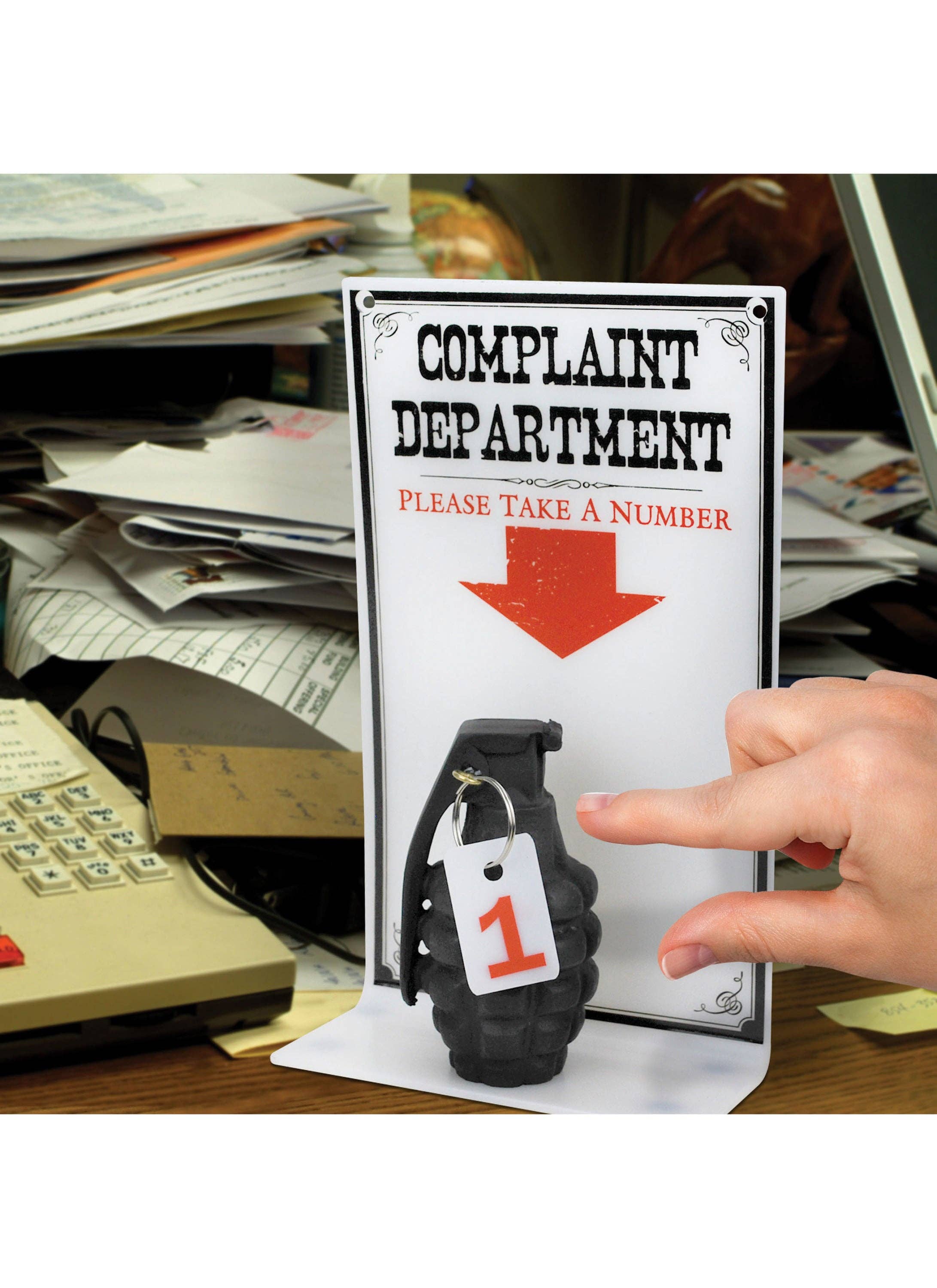 A hand reaches for a number tag attached to a grenade beneath a sign reading "Complaint Department, Please Take a Number," set on a cluttered desk.