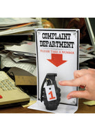 A hand reaches for a number tag attached to a grenade beneath a sign reading "Complaint Department, Please Take a Number," set on a cluttered desk.