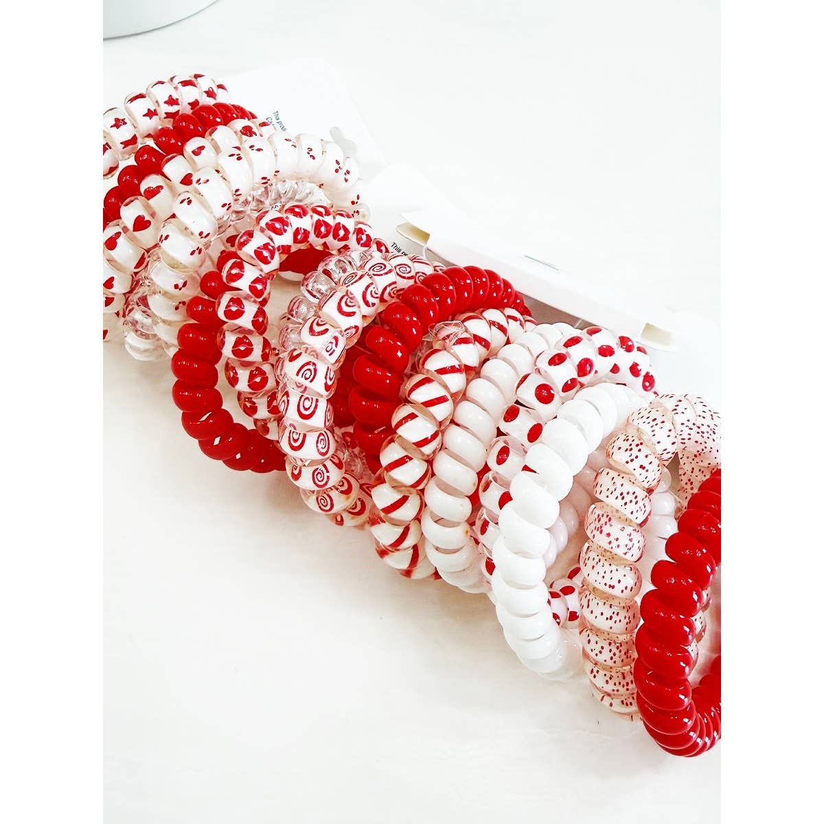 A collection of spiral hair ties in red, white, and mixed patterns, with heart and swirl designs, lies in a tidy row on a white surface.