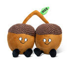 Two brown acorn plushies with smiley faces, their stems touching. A green embroidered leaf connecting them features the word 'deez' in black text. Both acorns have black legs sticking out from the bottom.