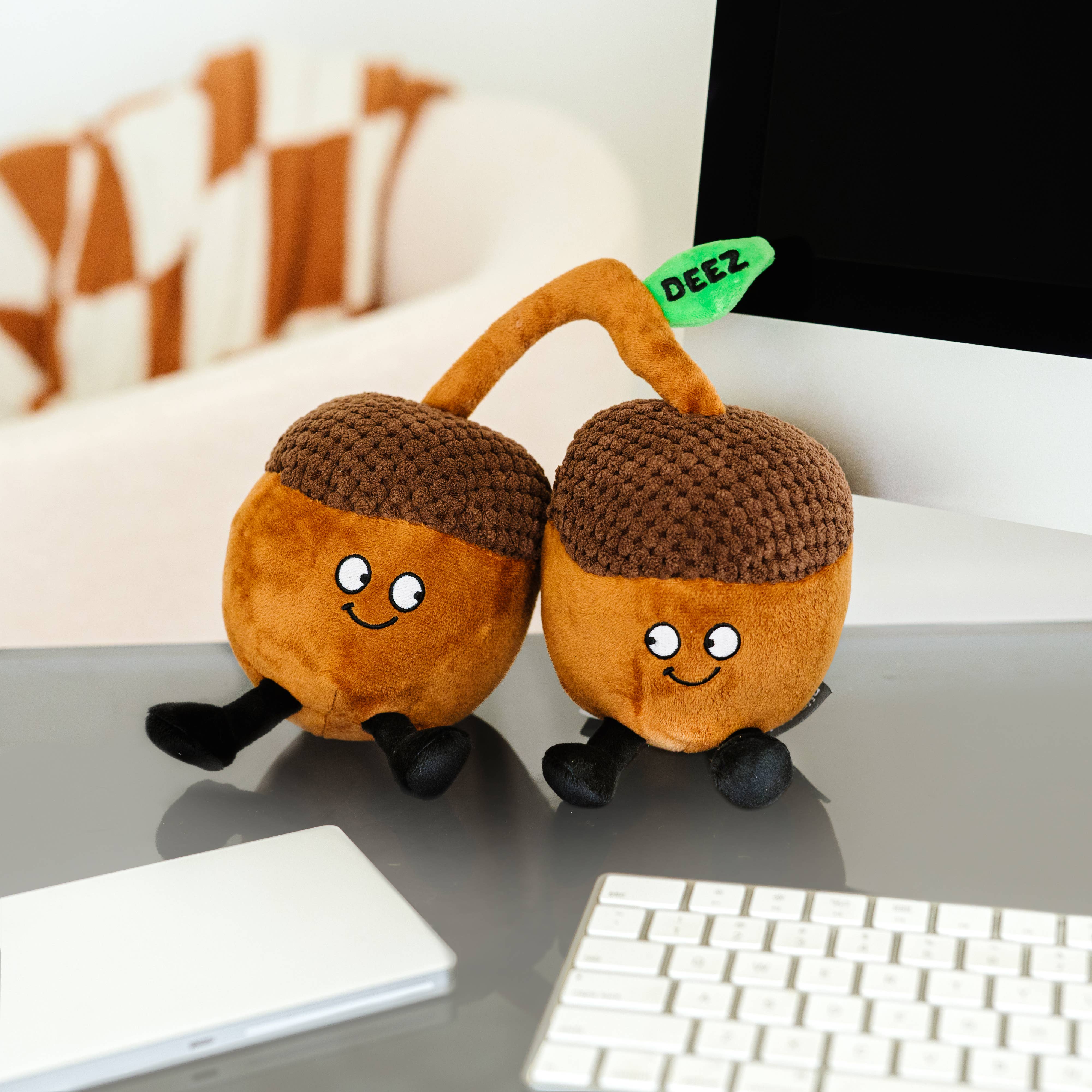 Two brown acorn plushies with smiley faces, their stems touching. A green embroidered leaf connecting them features the word 'deez' in black text. Both acorns have black legs sticking out from the bottom.