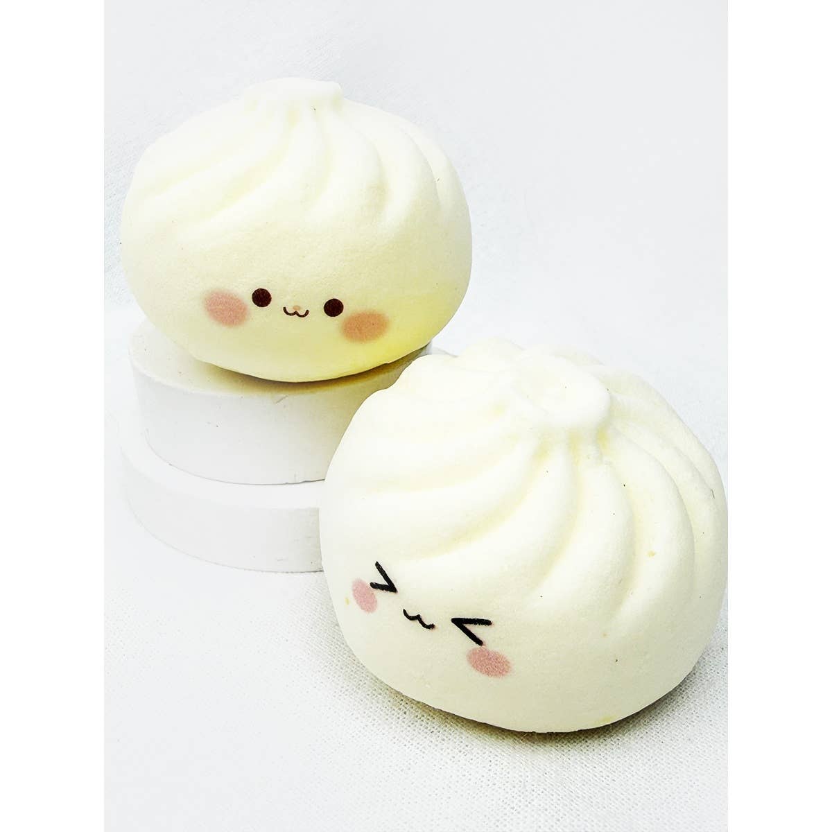 Two off-white dumpling-shaped slow-rising squishy toys with cute, simple anime-style smiley faces, staged on small white display blocks.