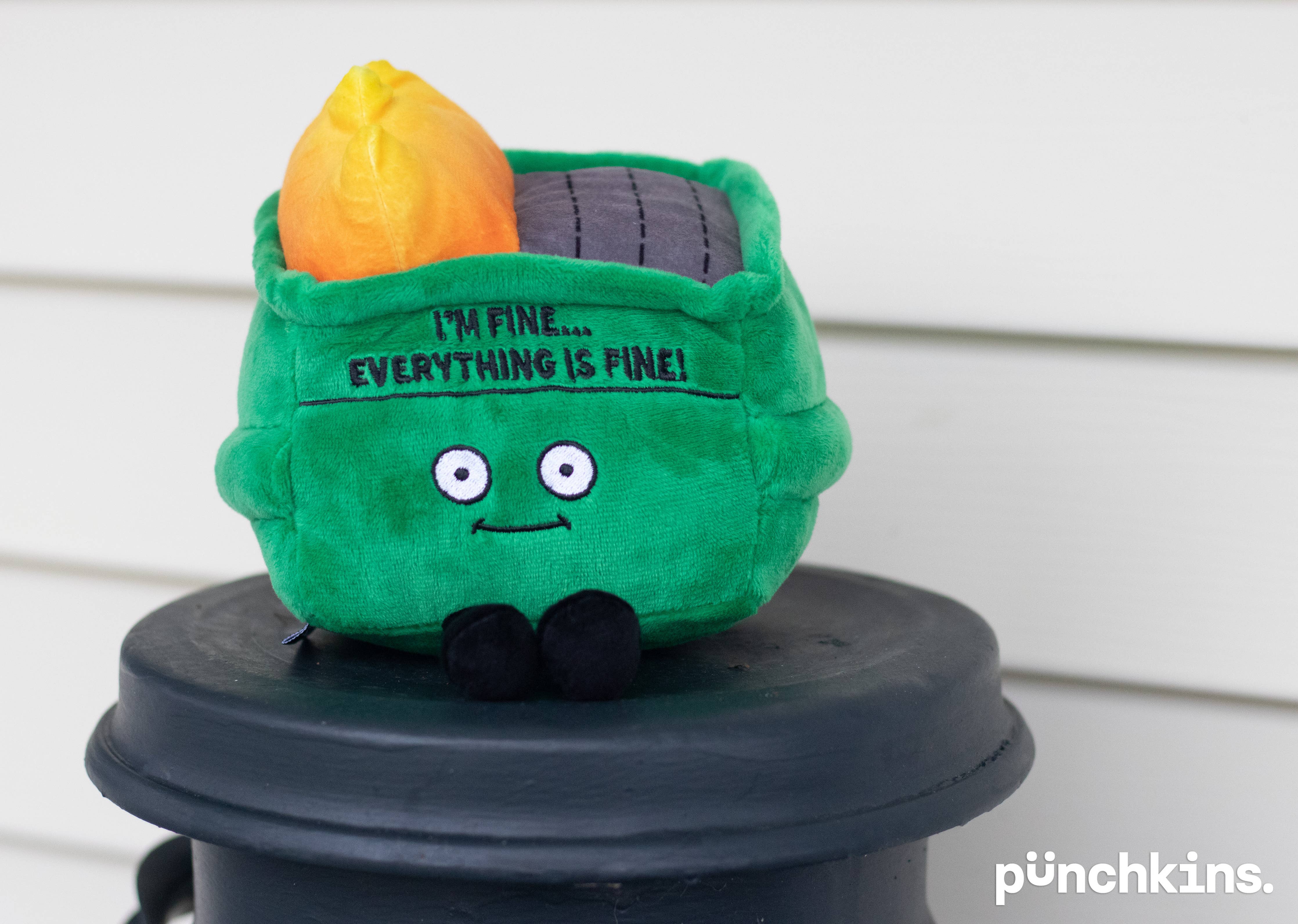 A plushie green dumpster fire with a smiley face, black embroidery that says 'i'm fine... everything is fine!' and black legs sticking out from the bottom. Yellow flames are coming out from the top."