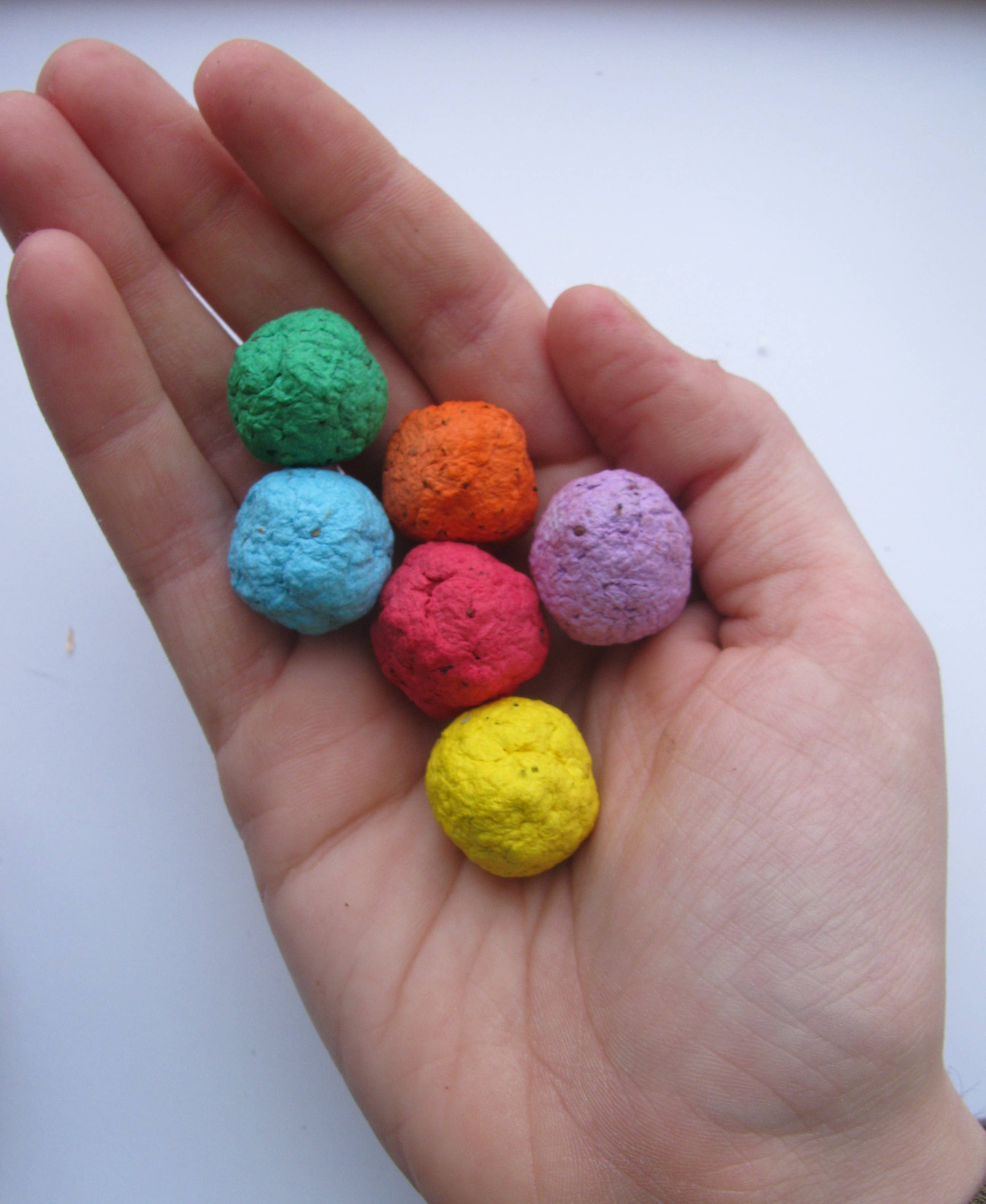 A hand holds six small, round seed bombs, each a different color: green, orange, blue, purple, red, and yellow, on a white background. Colorful and inviting.