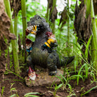 A dinosaur figurine playfully holds garden gnomes among lush green plants and soil, creating a whimsical and humorous garden scene.