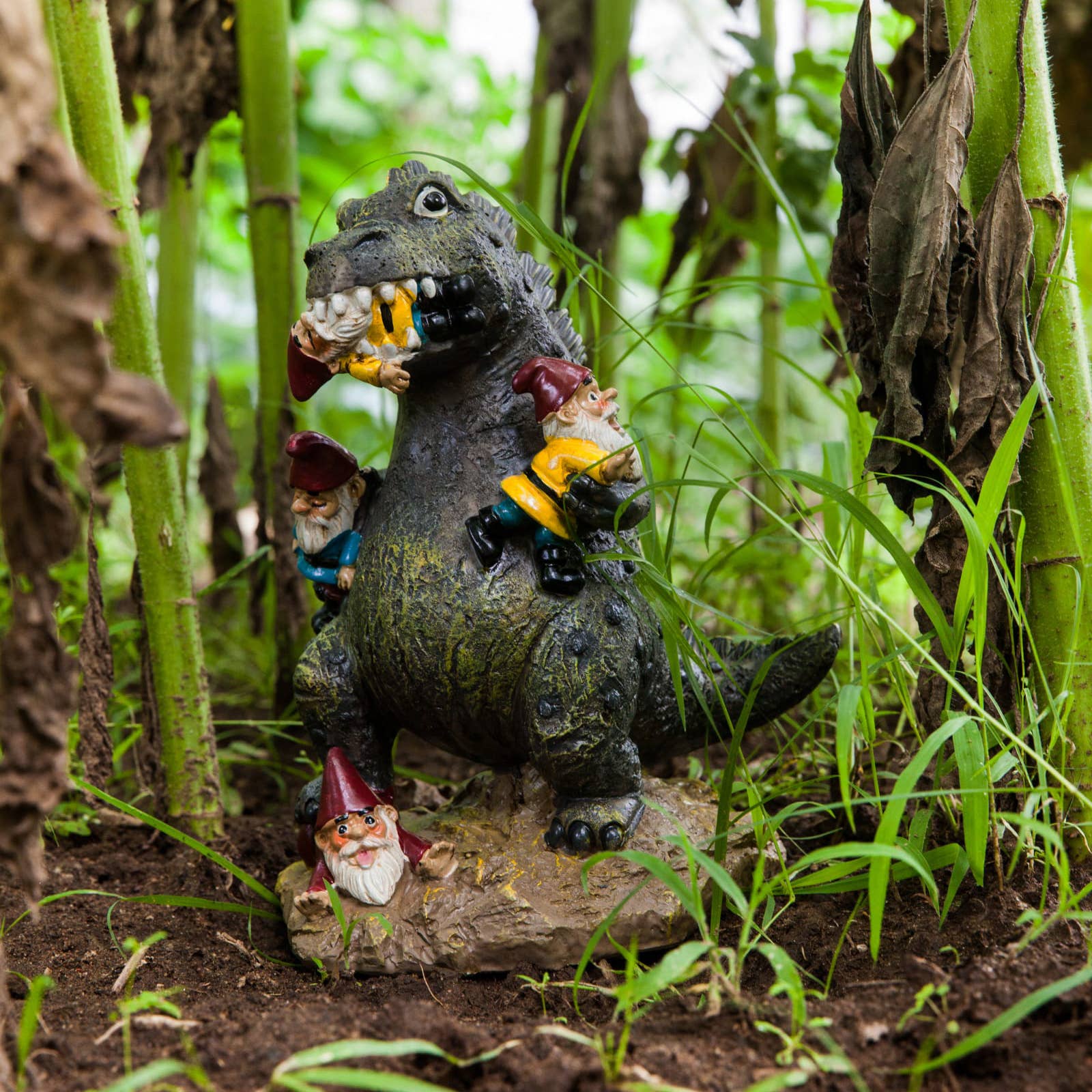 A dinosaur figurine playfully holds garden gnomes among lush green plants and soil, creating a whimsical and humorous garden scene.