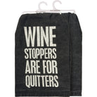 Dark kitchen towel with bold white text saying "Wine stoppers are for quitters." Humorous tone, displayed folded, hanging on dual hooks.