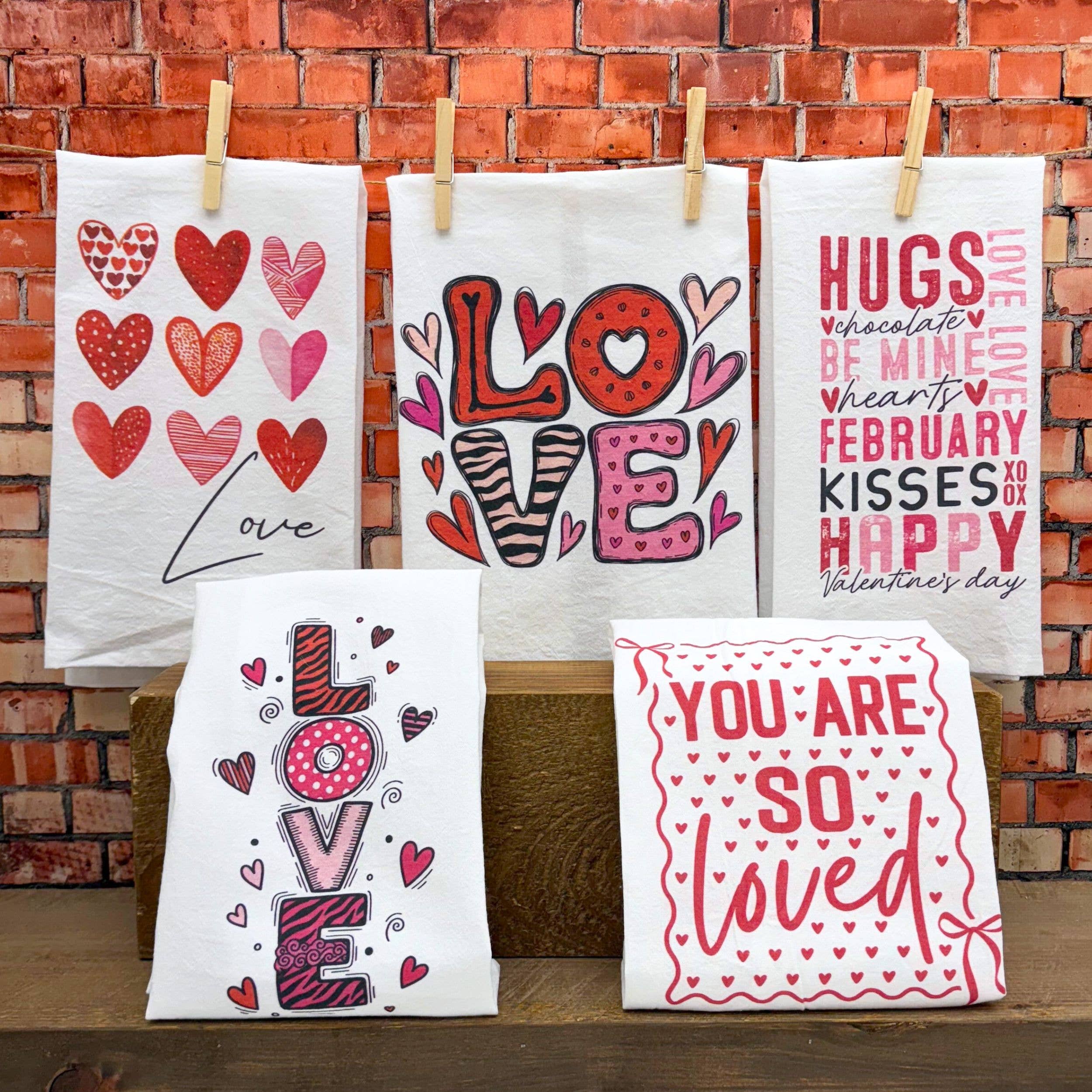 Valentine's-themed towels hang against a brick wall, featuring red and pink hearts, "LOVE" designs, and phrases like "You Are So Loved" in playful fonts.