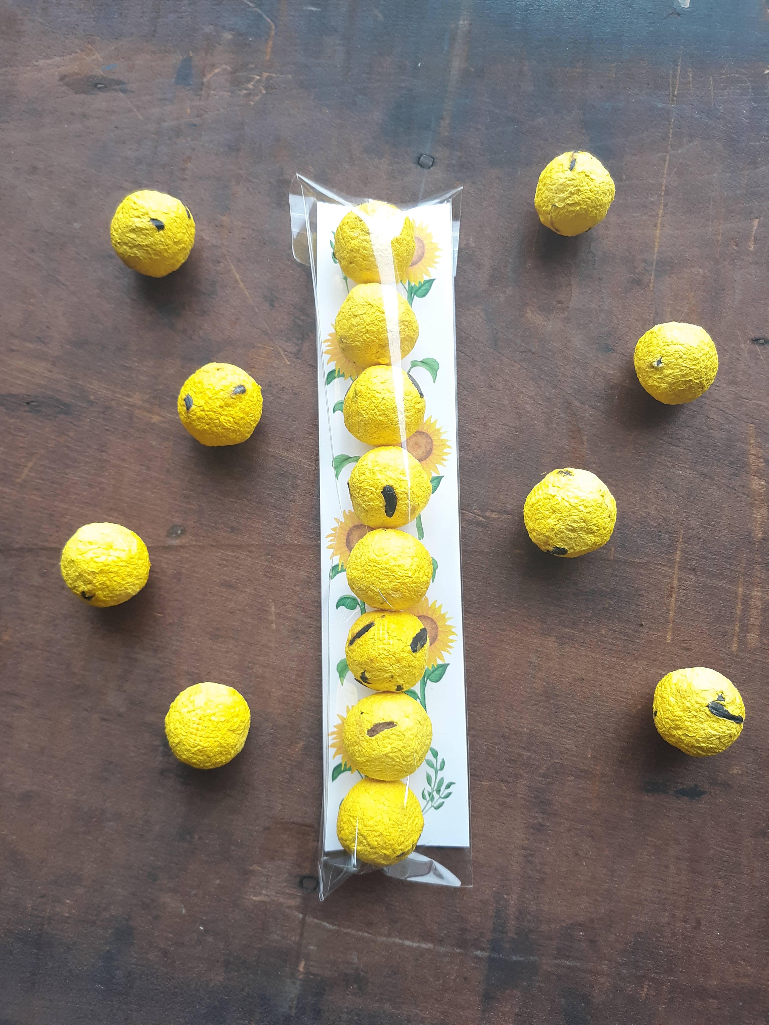 Yellow textured balls, some with black markings, are scattered on a wooden surface. A clear packet with a sunflower design holds similar balls.
