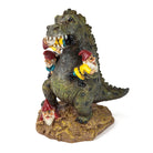 A whimsical garden sculpture depicts a green dinosaur playfully interacting with four garden gnomes. The gnomes appear to be climbing and clinging to the dinosaur, with one gnome in the dinosaur's mouth, creating a humorous and mischievous tone.