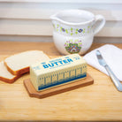 Stick of salted butter on a wooden tray beside sliced bread, a floral ceramic jug, a butter knife, and a white napkin. Cozy kitchen setting.