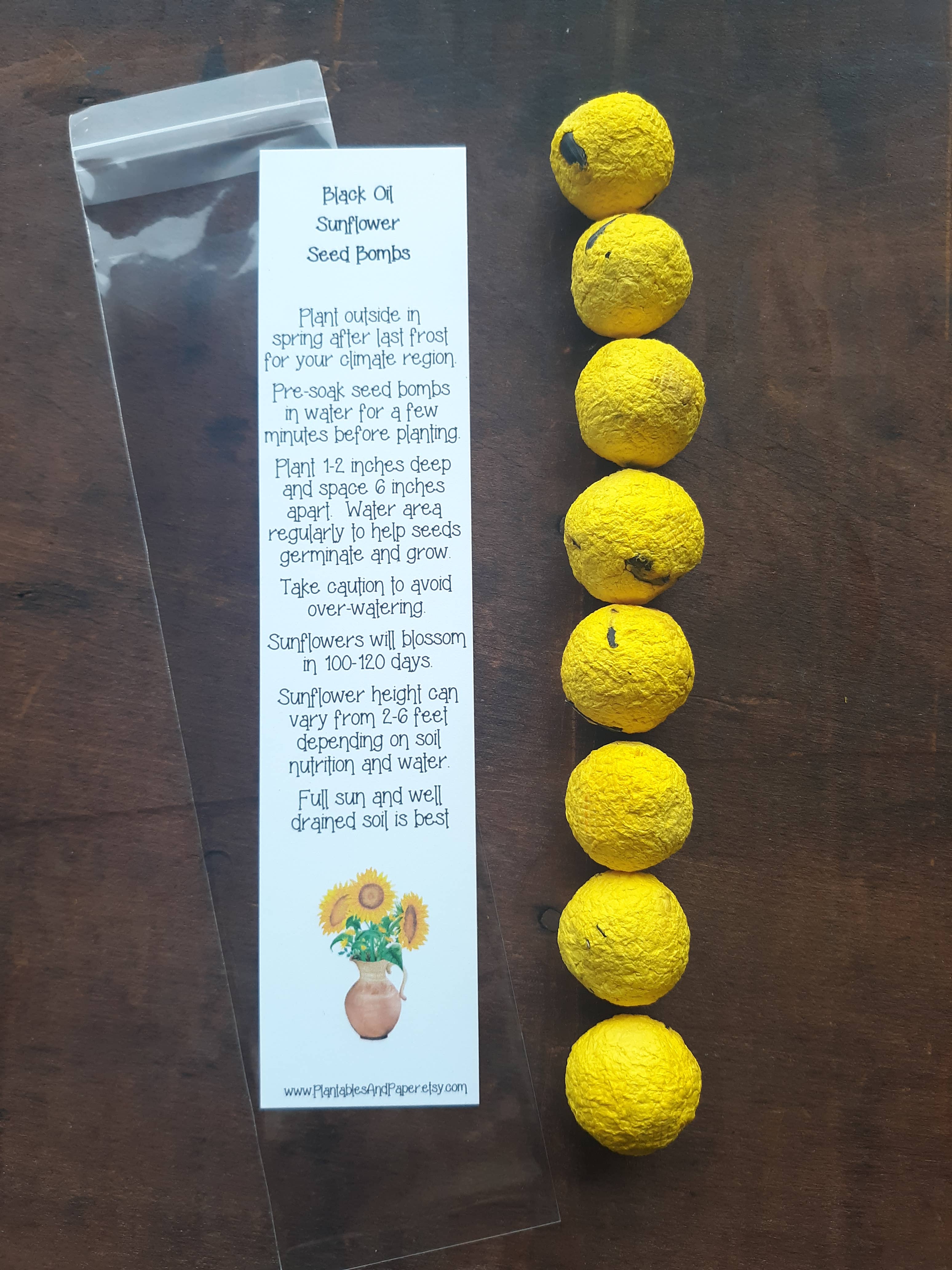 Seven yellow sunflower seed bombs are aligned vertically on a wooden surface. Beside them is a white instruction leaflet detailing planting and care tips for the seed bombs. The leaflet includes a small image of sunflowers in a terracotta pot.