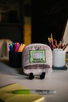 A gray computer plush with a winky face, featuring a green Excel-style sheet embroidered with the words "Freak in the Sheets" in black. It also has an outlined keyboard design and black legs sticking out from the bottom. Plush is sitting on a desk.