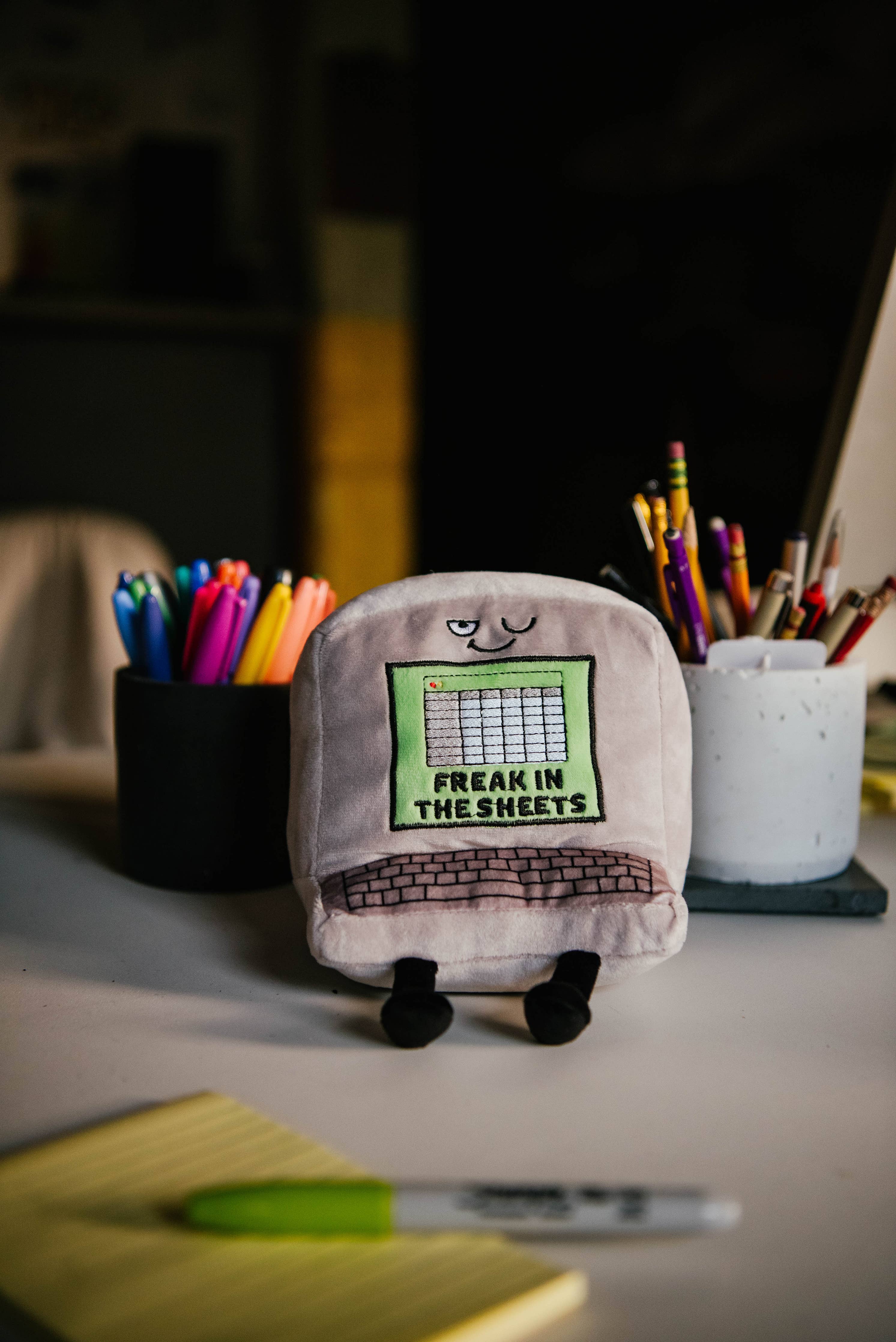 A gray computer plush with a winky face, featuring a green Excel-style sheet embroidered with the words "Freak in the Sheets" in black. It also has an outlined keyboard design and black legs sticking out from the bottom. Plush is sitting on a desk.