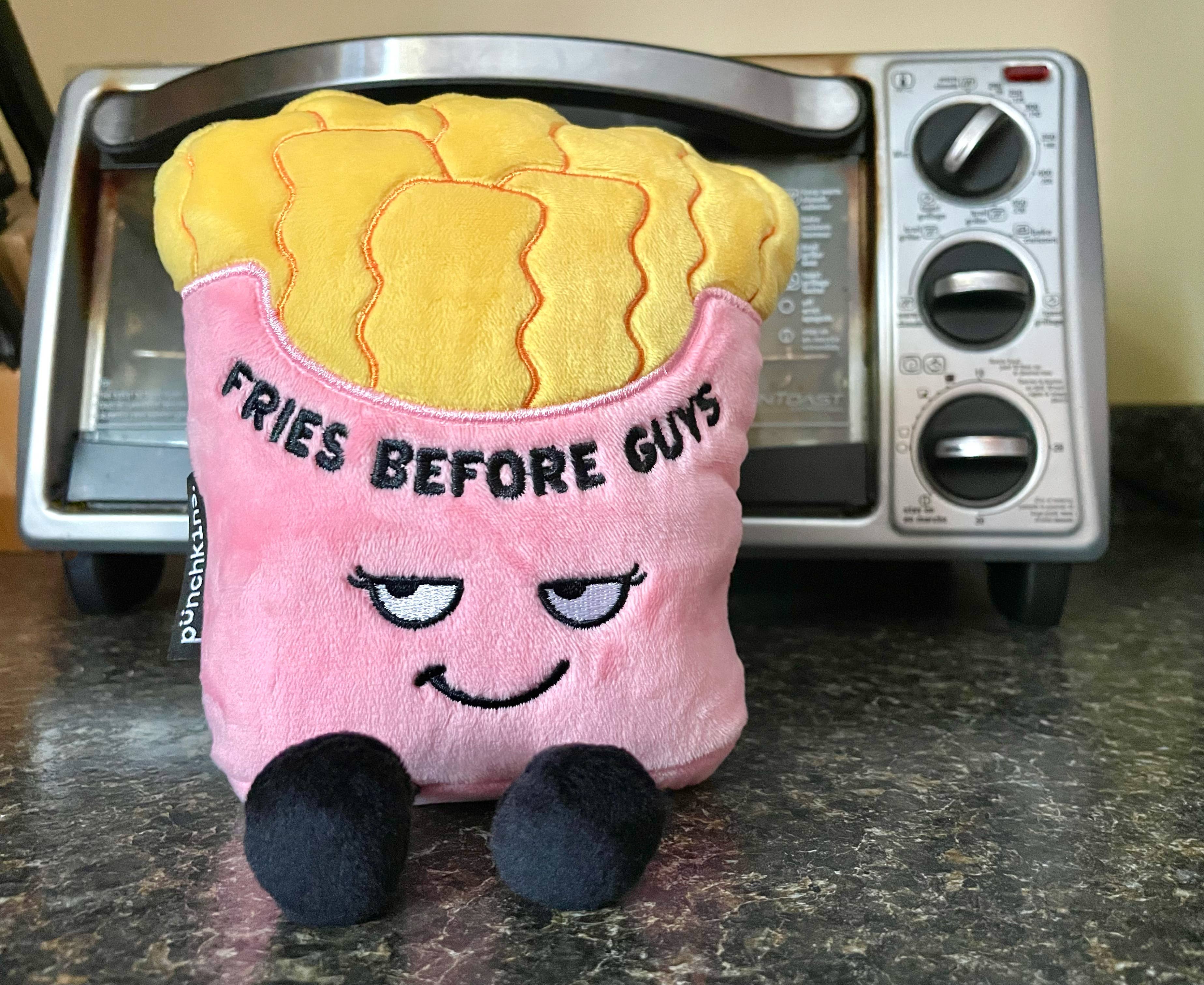 A pink fries container holding yellow fries. The pink section has a sly smiley face and black text that says "Fries before guys." The plushie has black legs sticking out.