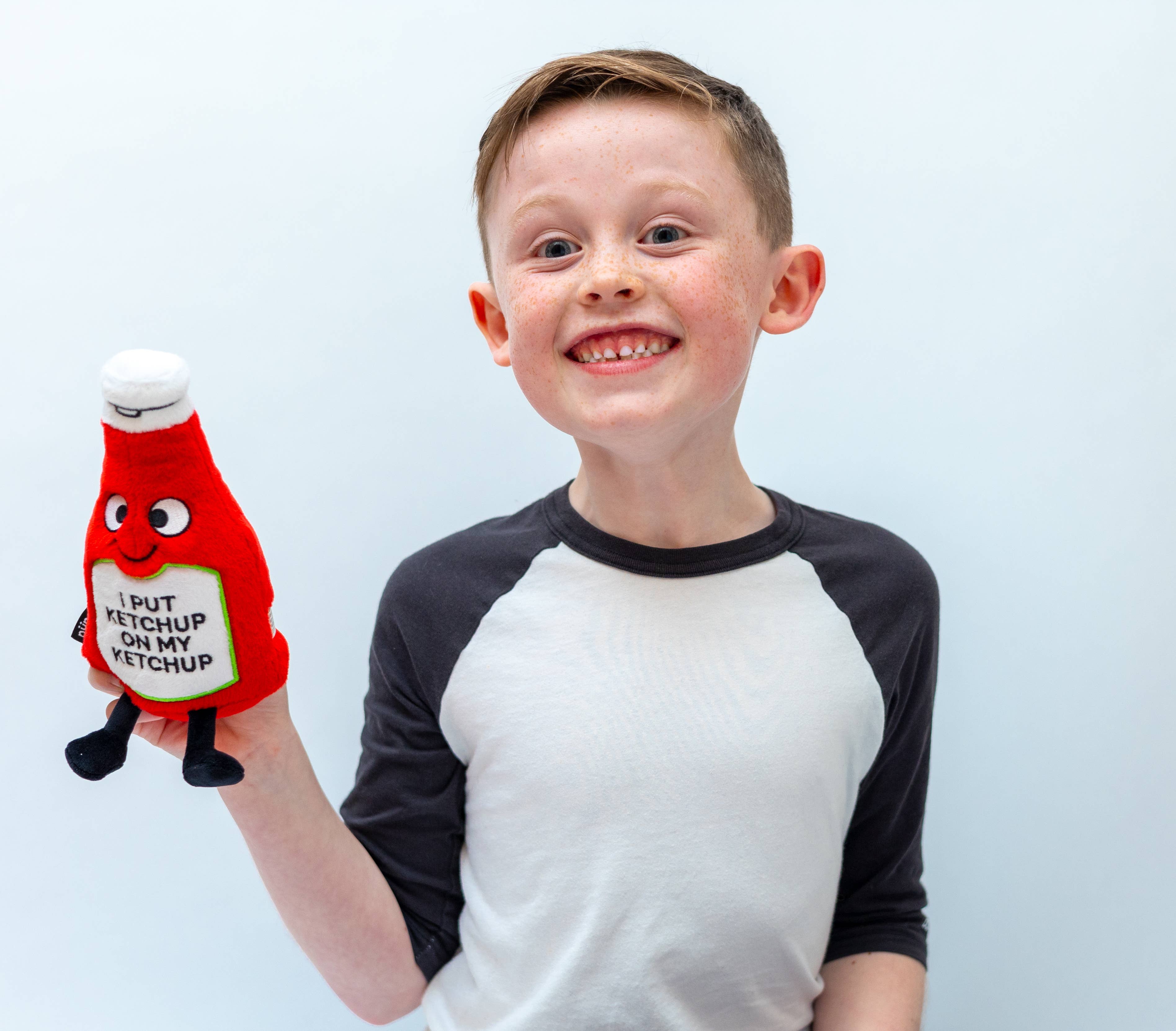 Child holding a ketchup bottle plushie with a smiley face and a white belly featuring black text that says "I put ketchup on my ketchup." It has a white lid and black legs sticking out.