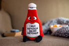 A ketchup bottle plushie with a smiley face and a white belly featuring black text that says "I put ketchup on my ketchup." It has a white lid and black legs sticking out.