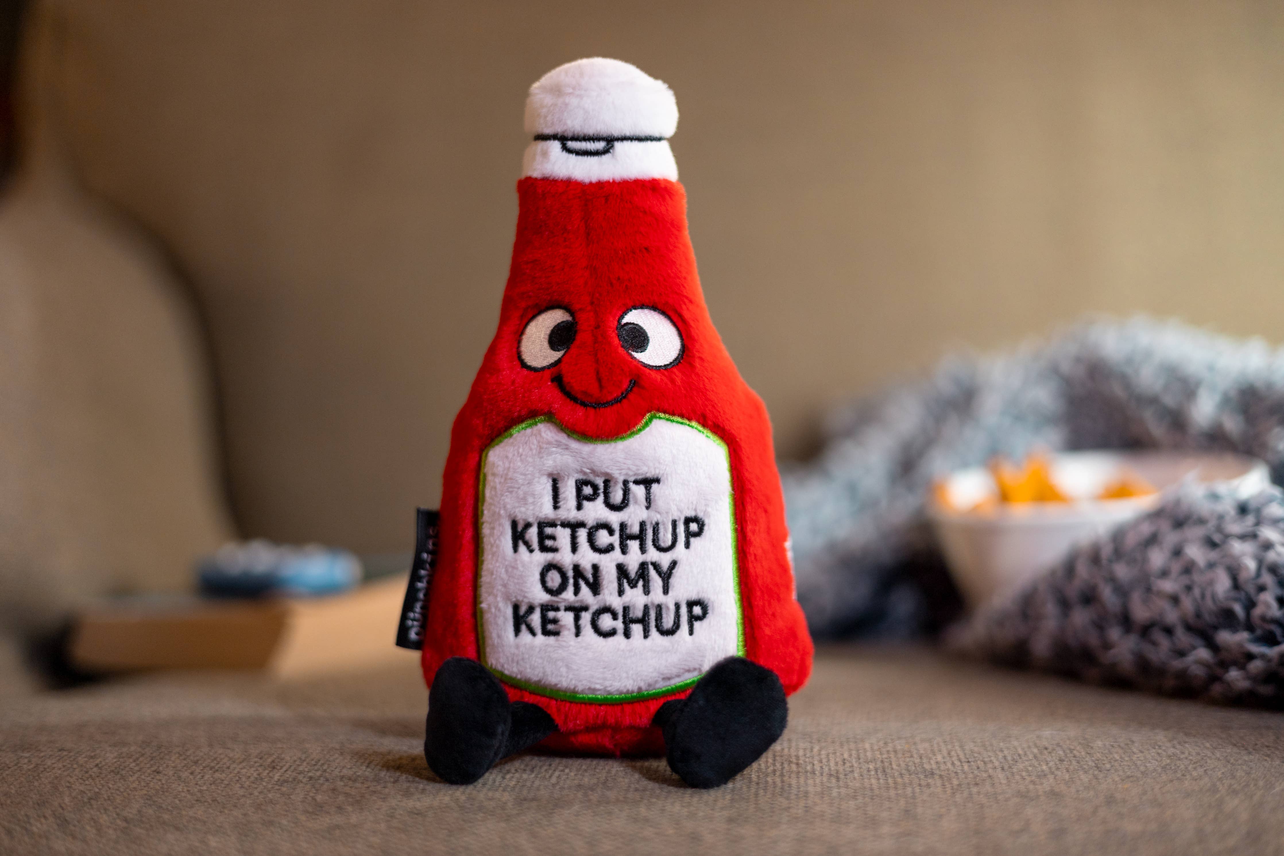 A ketchup bottle plushie with a smiley face and a white belly featuring black text that says "I put ketchup on my ketchup." It has a white lid and black legs sticking out.