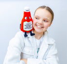 Child holding a ketchup bottle plushie with a smiley face and a white belly featuring black text that says "I put ketchup on my ketchup." It has a white lid and black legs sticking out.