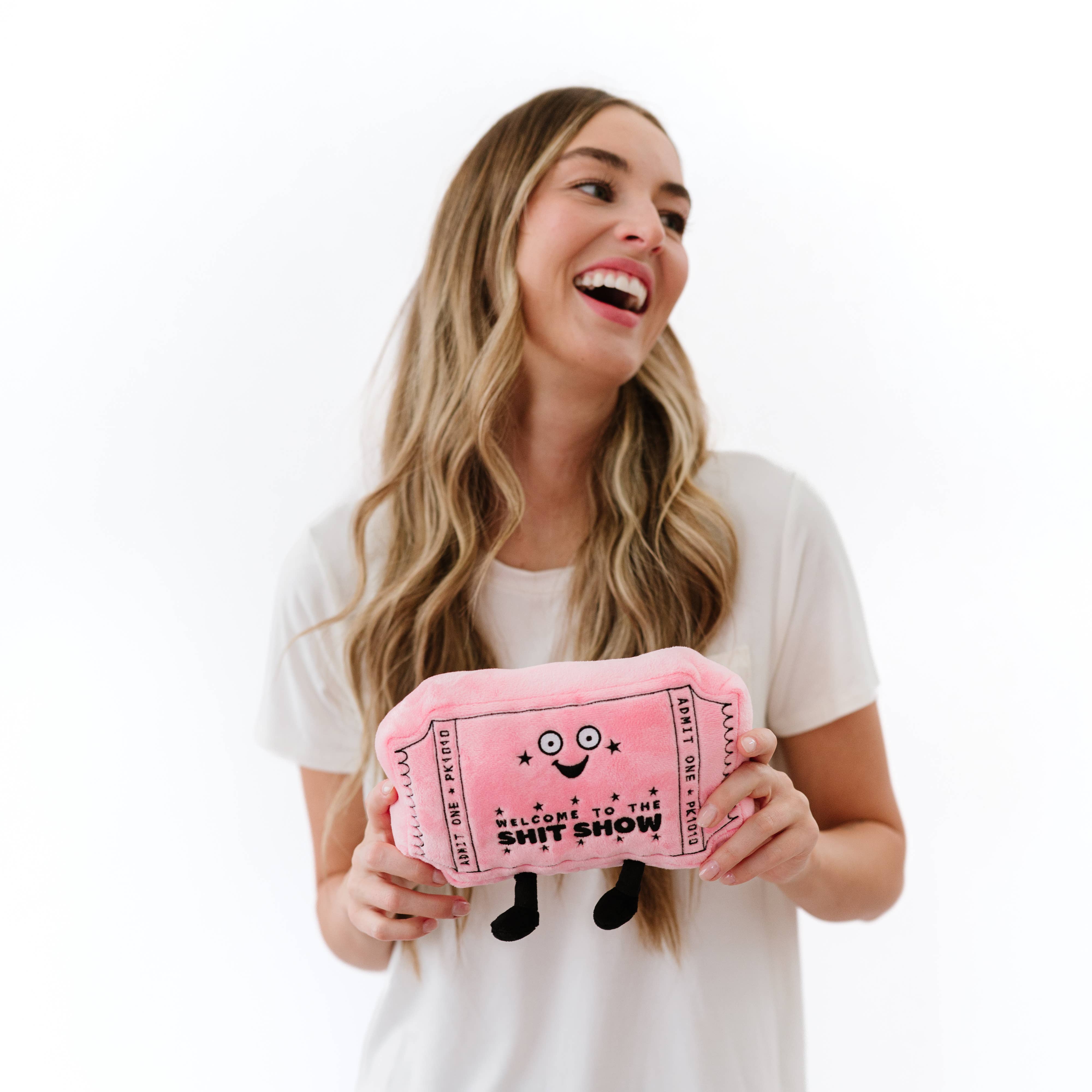 Woman laughing while holding A pink plushie shaped like a movie ticket with a smiley face. It features black embroidery with the words "welcome to the shit show.